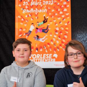 Luis und Konrad sitzen als Sieger vor dem Plakat zum Bezirksentscheid und freuen sich.