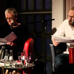 Die WDR-Sprecher*in Regina Münch und Martin Groß sitzen auf dem roten Sofa und lesen Texte vor. - Foto: Judith Mengel