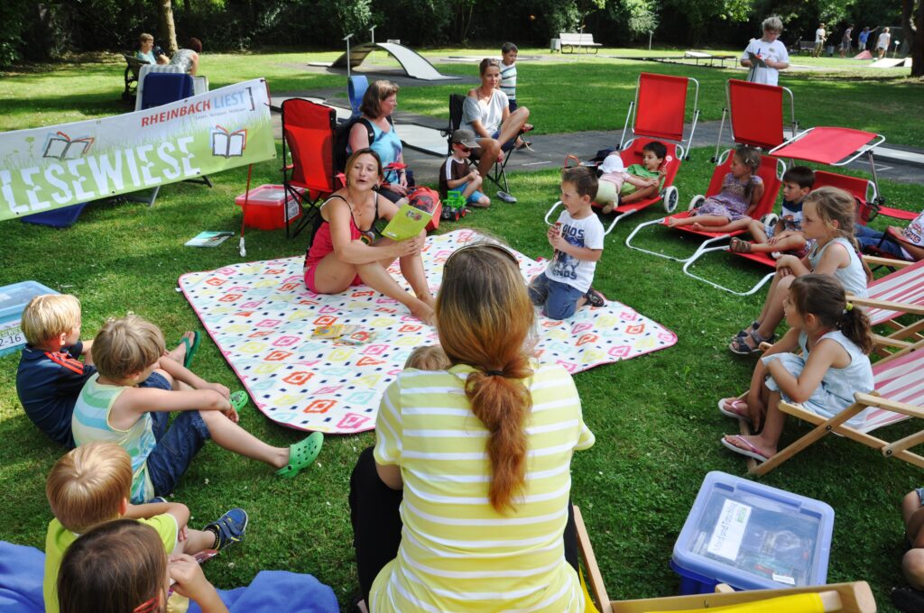 Auf der Picknick-Decke liest die Vorleserin bei strahlenden Sonnenschein den anwesenden Kindern vor.