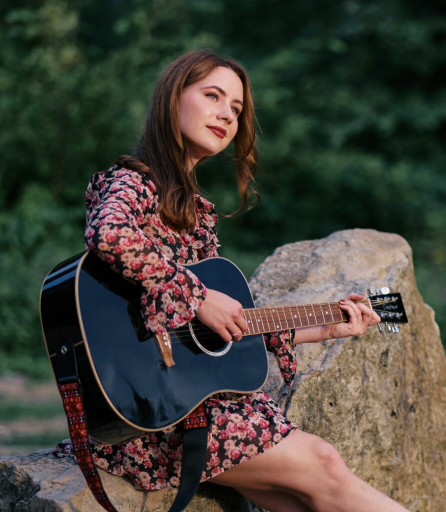 Annalena Sybon sitzt mit einer klassischen Gitarre auf einem Holzstamm und singt. Foto F. Siepen