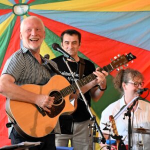 Dieter Peters an der Gitarre singend und lachend mit Bandmitgliedern im Rücken