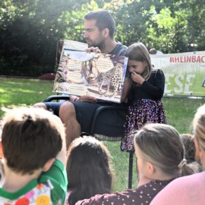 Bürgermeister sitzt vor den Kindern udn zeigt die Bilder aus dem Bildebuch Bürgermeister liest aus dem Bilderbuch „Kleine Pandas, großes Versprechen“ und liest vor.